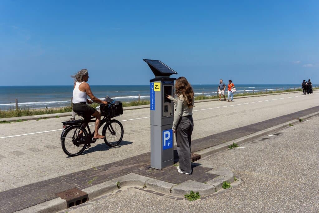 Person using a parking meter on Zandvoort boulevard while a cyclist passes by with the beach and sea in the background.
